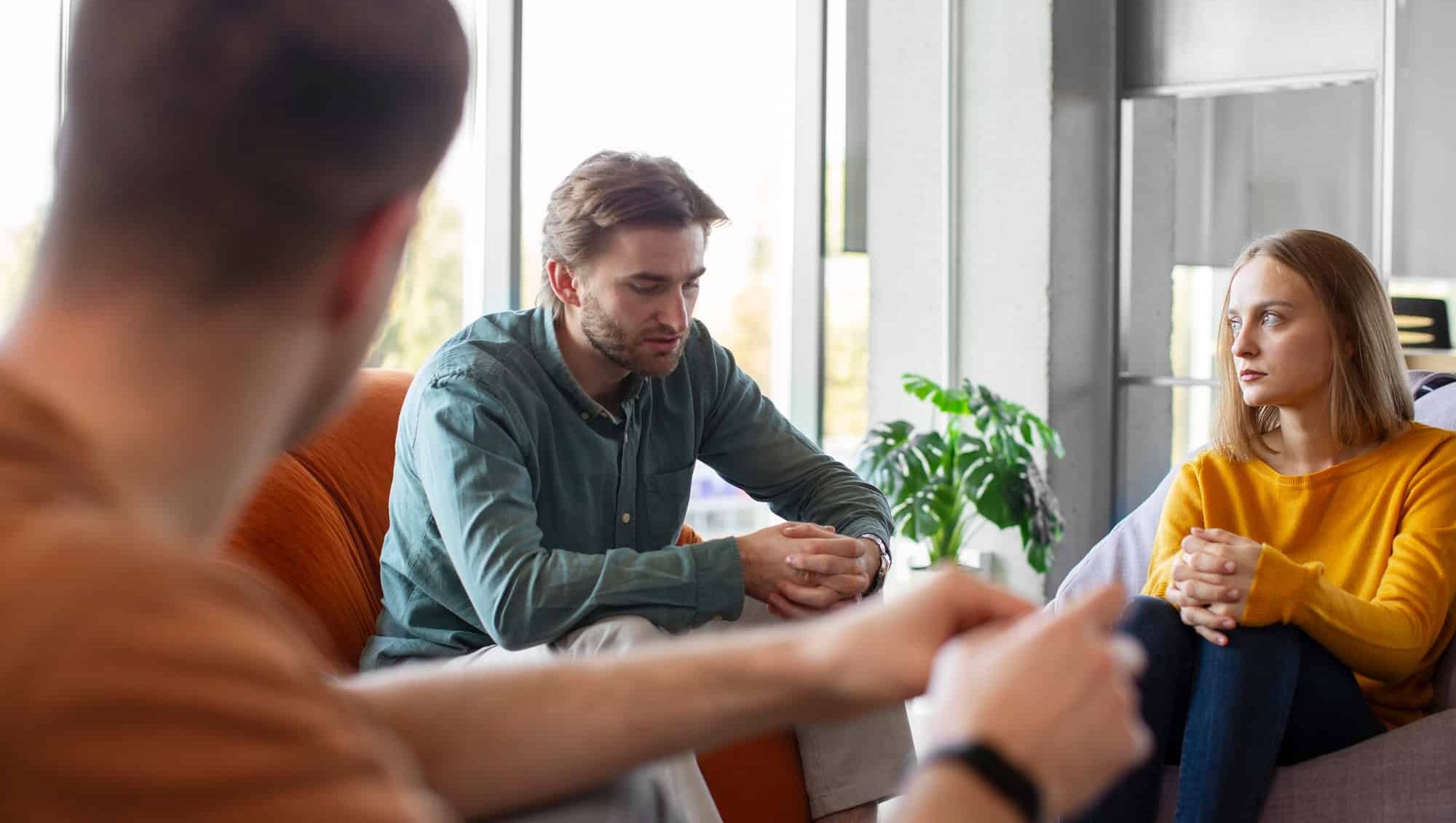 Tre personer sitter i samtale, to med alvorlige uttrykk under en gruppeterapitime.