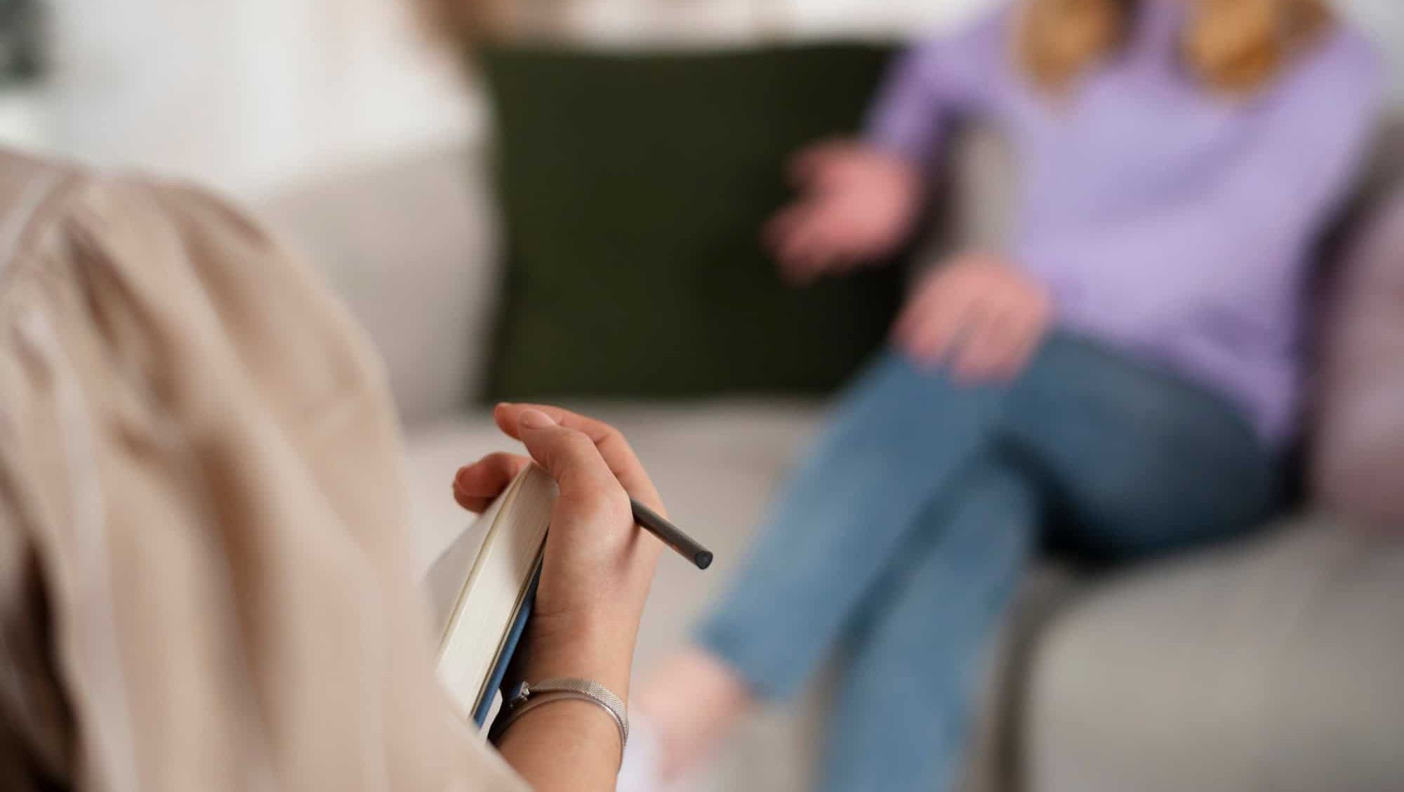 Terapeut noterer mens klient i lilla genser snakker på en sofa under samtaleterapi.